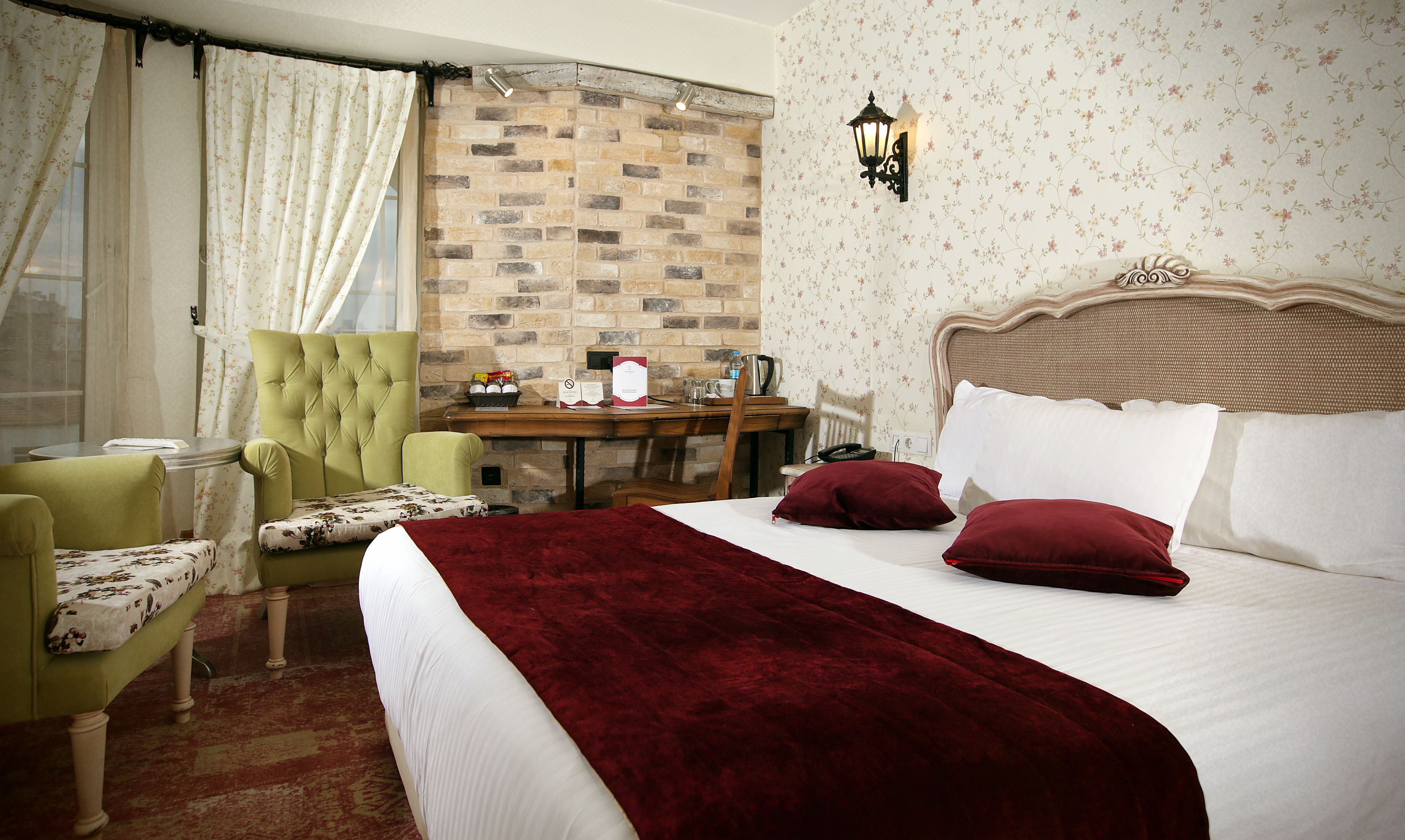 A hotel room with a bed, a green armchair, a wooden desk, and a brick wall.