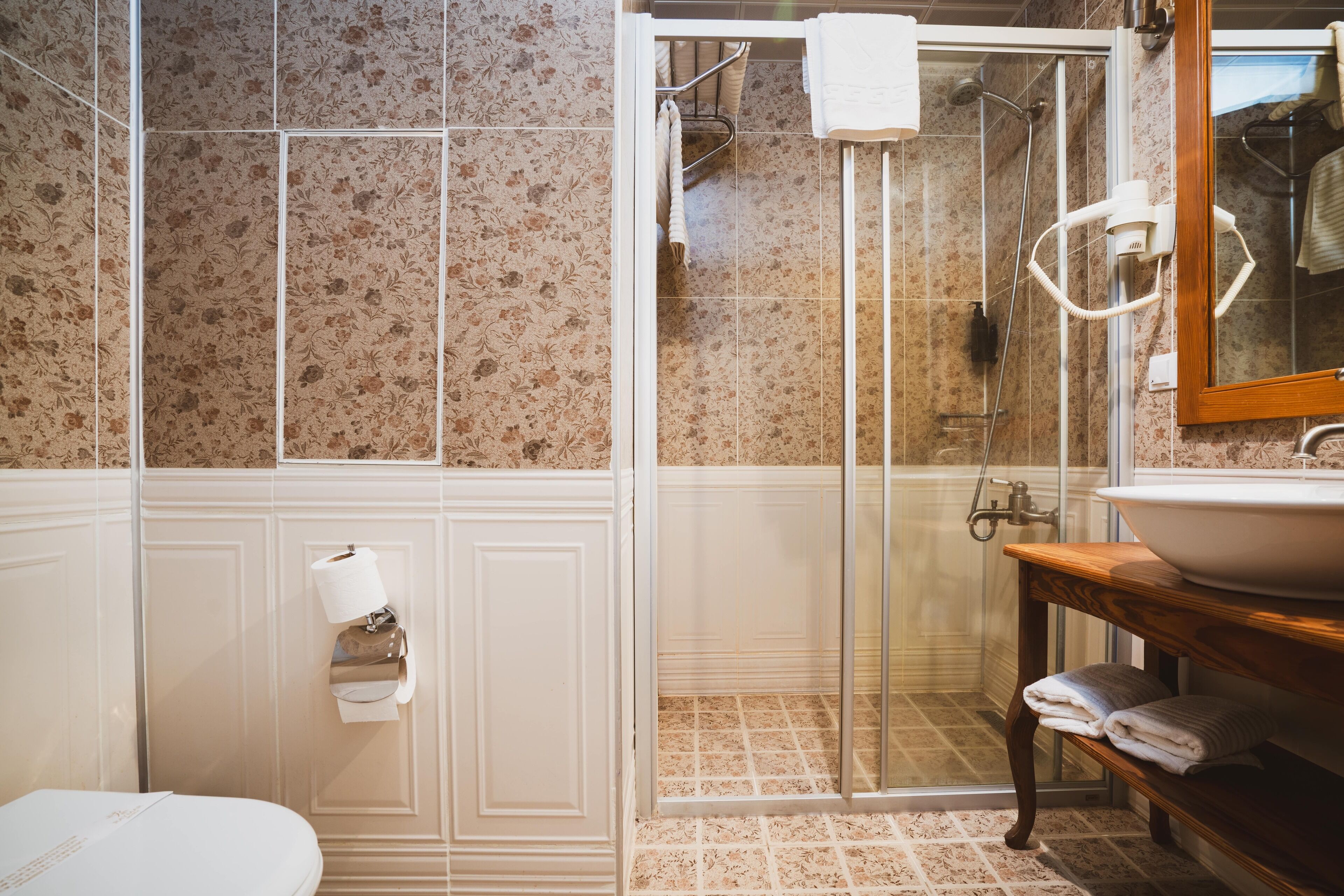 Un baño con ducha envuelta en vidrio, un lavamanos con tocador de madera y un inodoro con dispensador de toallas de papel.