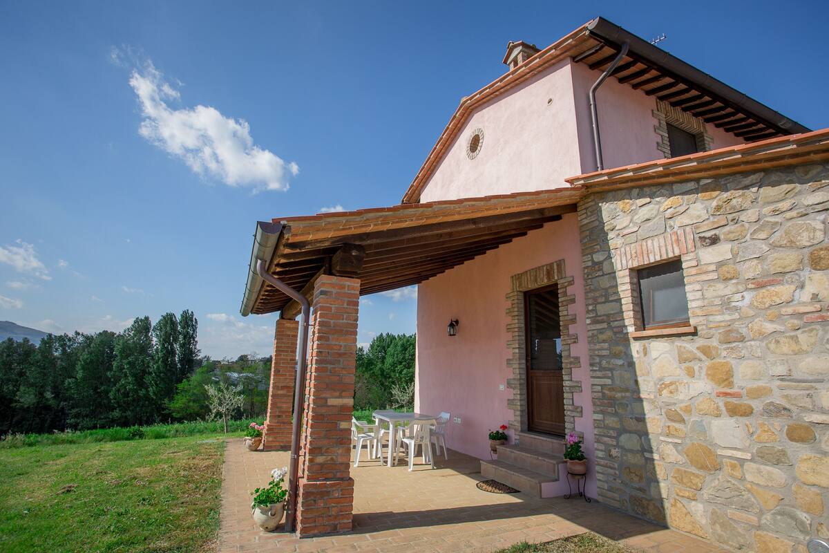 A stone house with a pink exterior, a covered patio with chairs, and a view of the surrounding greenery.
