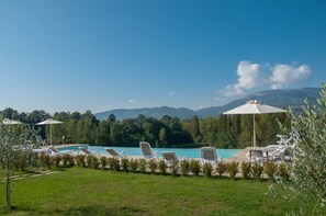 Zona de piscina con sillas de descanso y sombrillas, rodeada de vegetación y montañas al fondo.