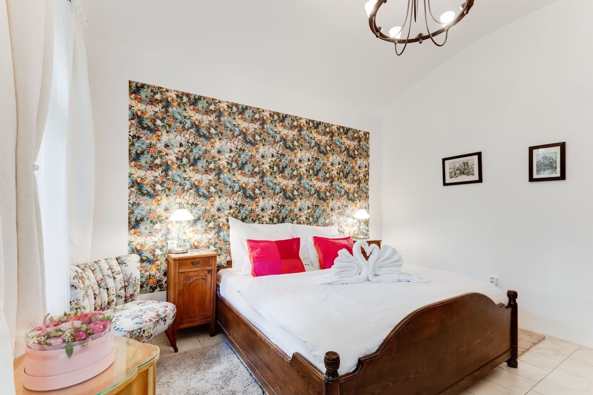Quarto com papel de parede floral, cama de madeira, duas luminárias de cabeceira e uma mesa pequena com um arranjo de flores.