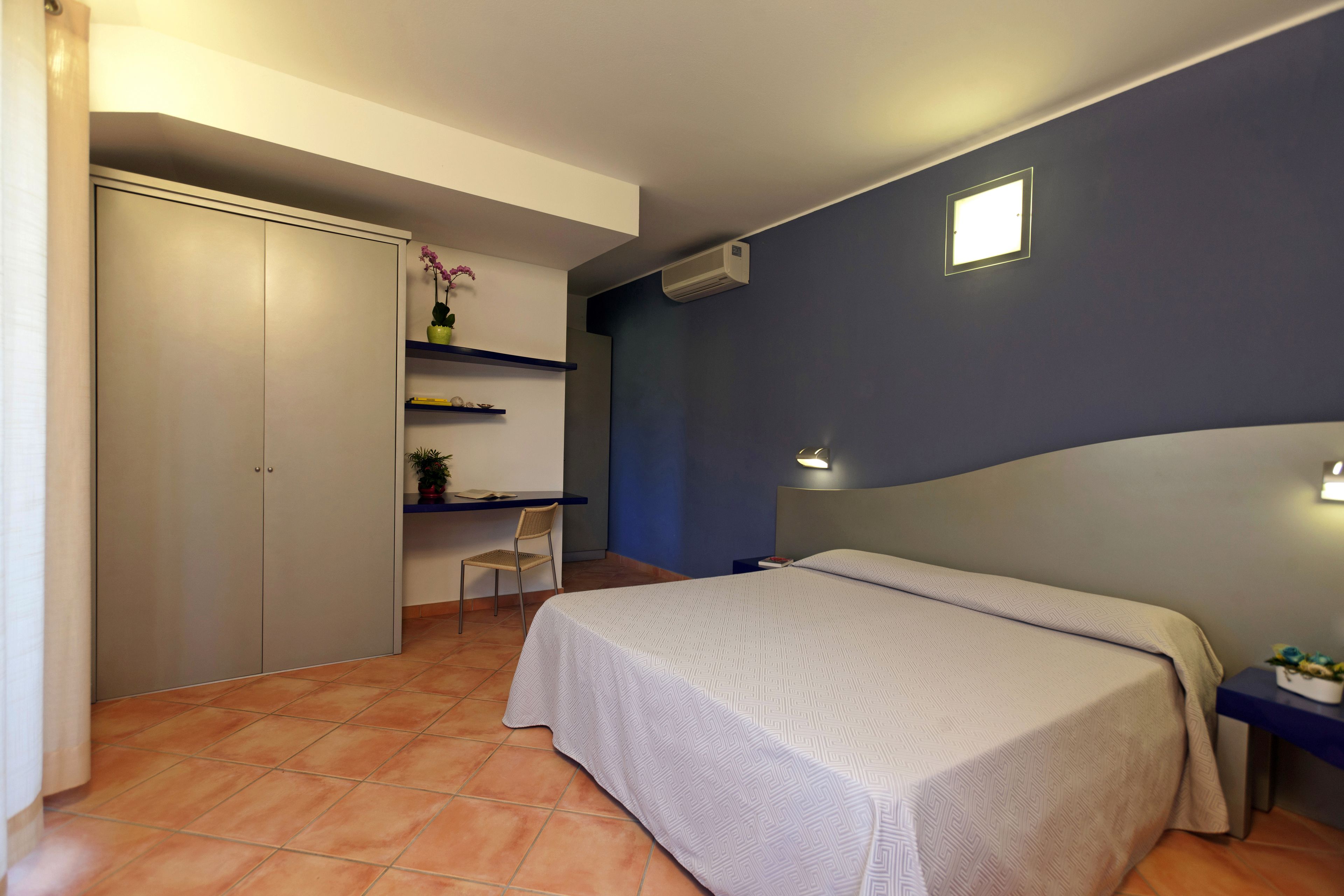 A bedroom with a bed, a desk, a chair, a wardrobe, and a small shelf with plants.