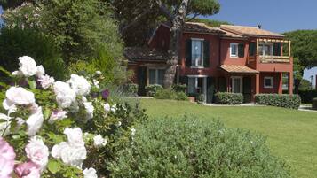 Una casa de dos pisos con fachada roja, un árbol grande en el frente y un jardín bien cuidado con flores blancas y rosadas.