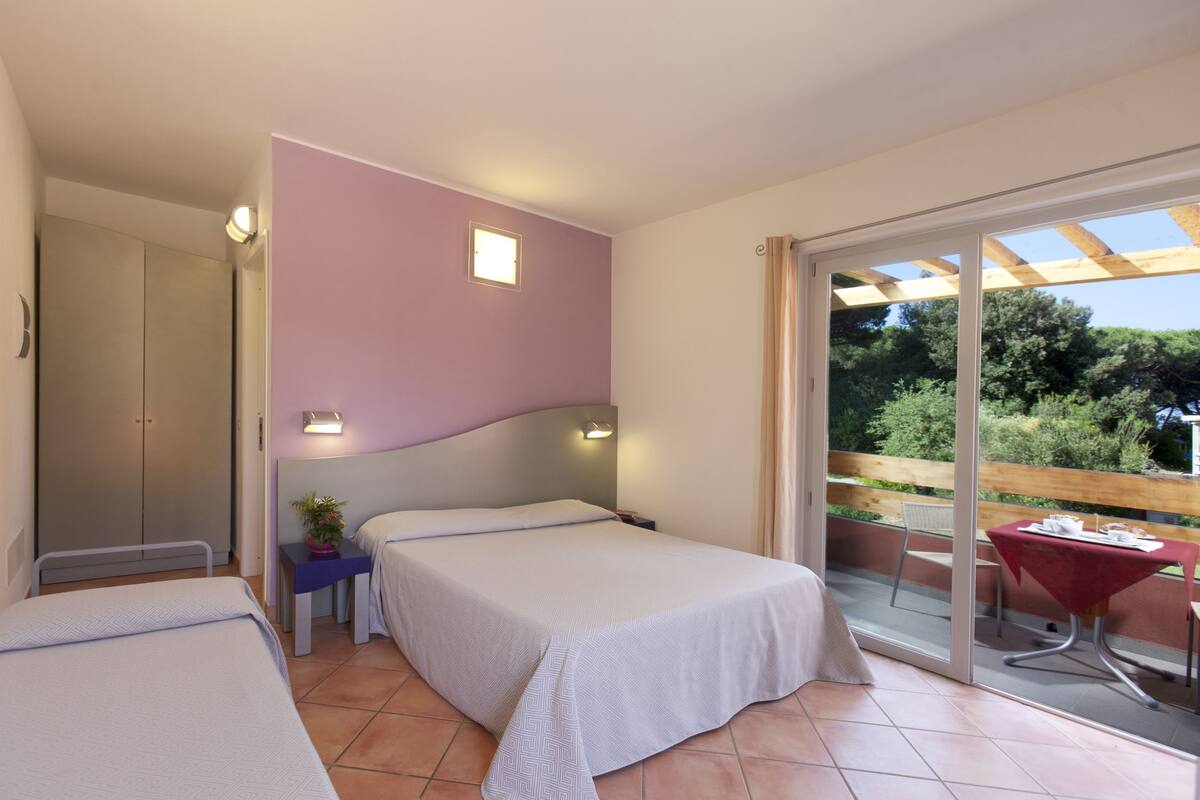 A bedroom with two beds, a small table with a plant, a wardrobe, and a balcony with a table and chair.