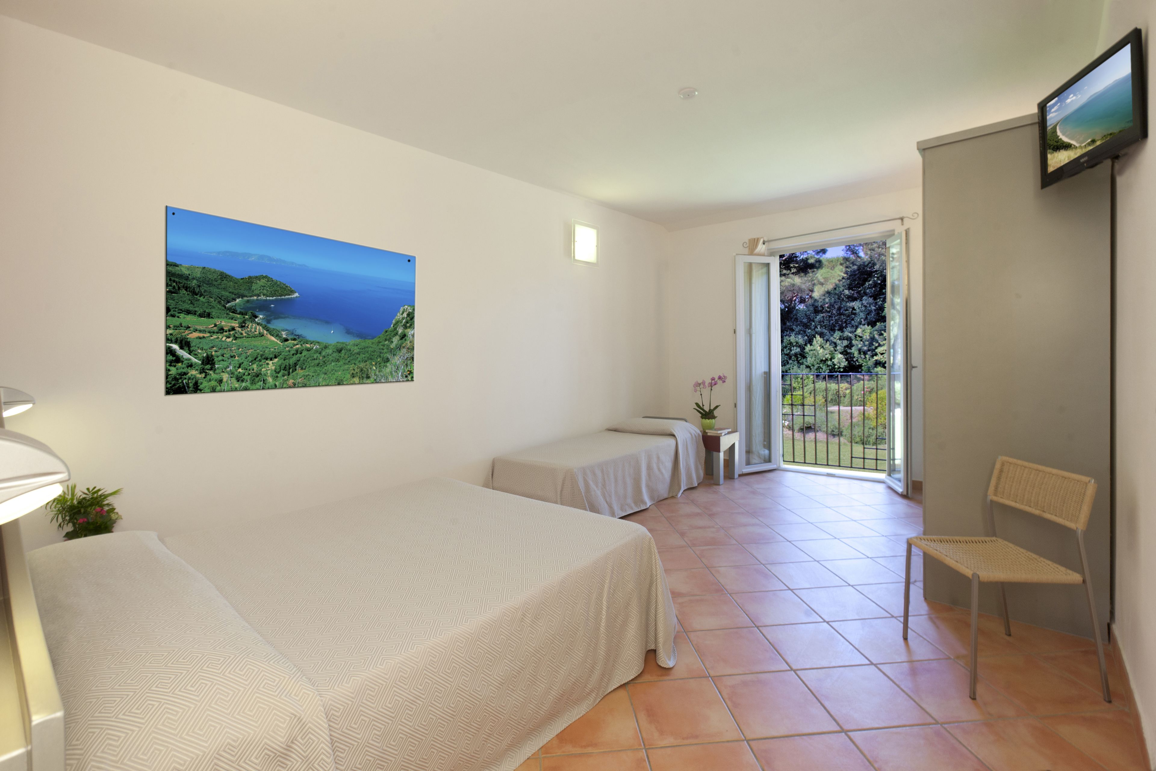 A bedroom with a bed, a small table with a plant, a chair, and a balcony with a view of greenery and a body of water.