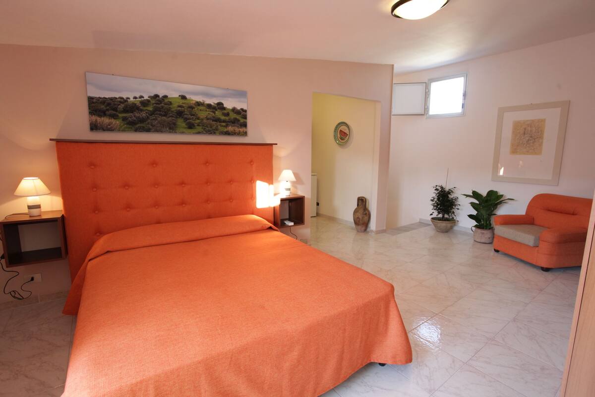 A bedroom with a large bed, a nightstand, a lamp, a painting, a door, a window, and a chair.