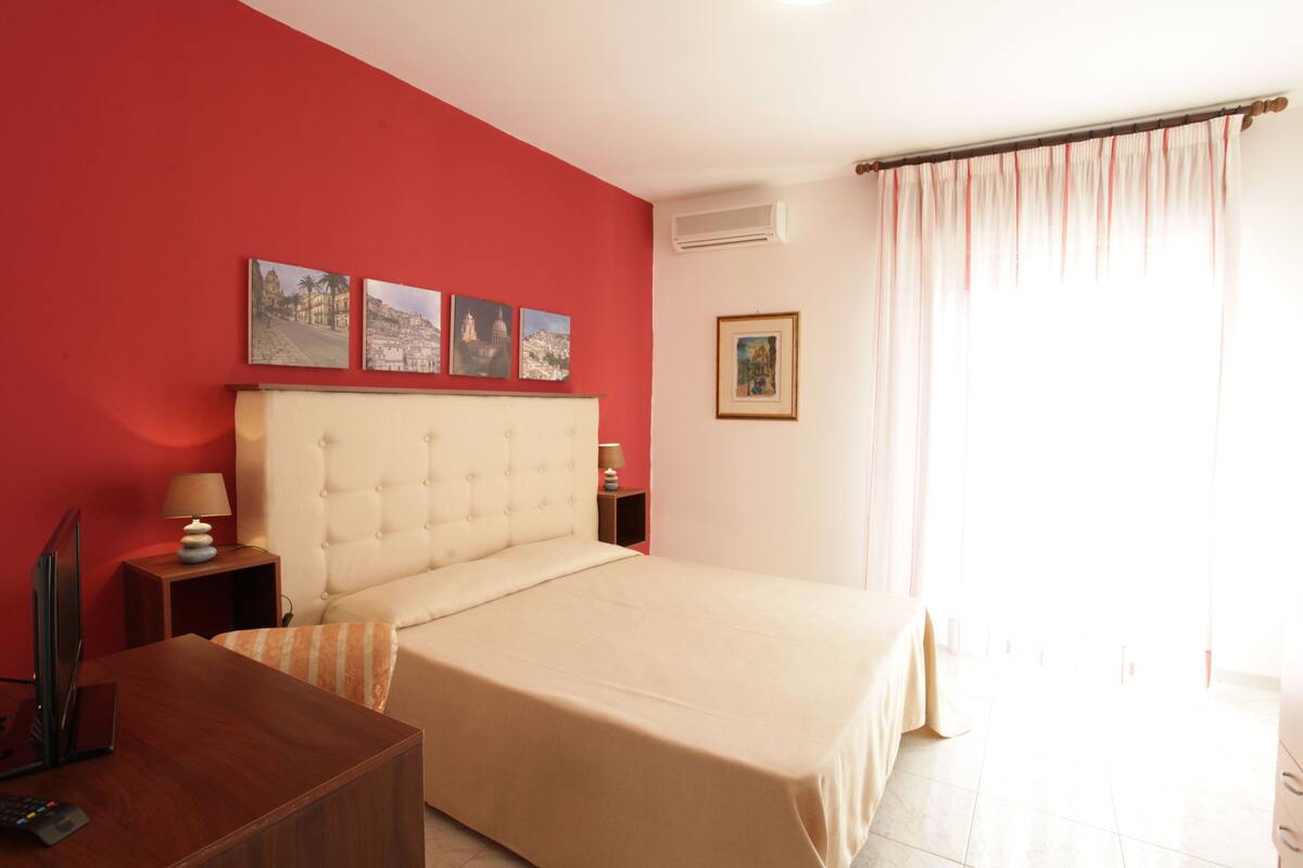 A hotel room with a large bed, a wooden desk, a red wall, and three framed pictures.