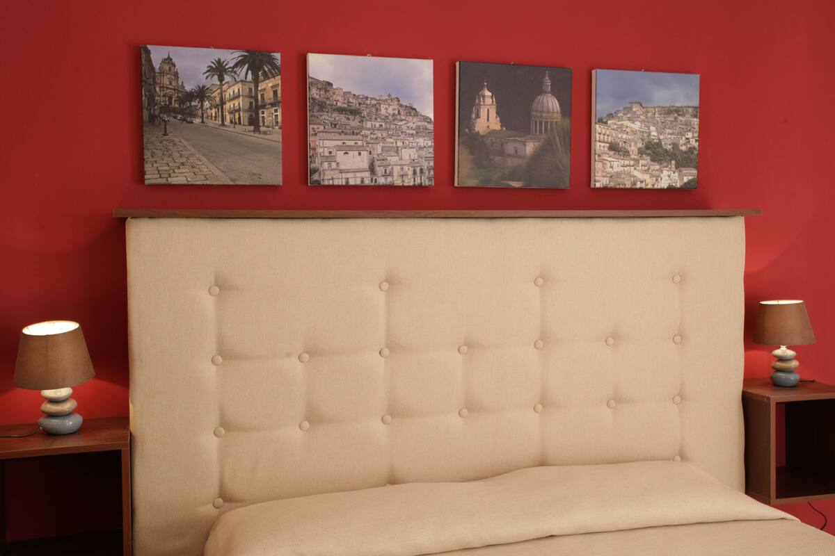 A bedroom with a tufted headboard, a bed, two bedside tables with lamps, and three framed cityscape pictures on the wall.