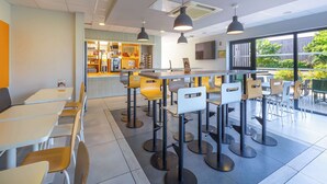 Een modern café met een bar, verschillende zitmogelijkheden zoals tafels en stoelen, en een groot raam met uitzicht op het buiten.