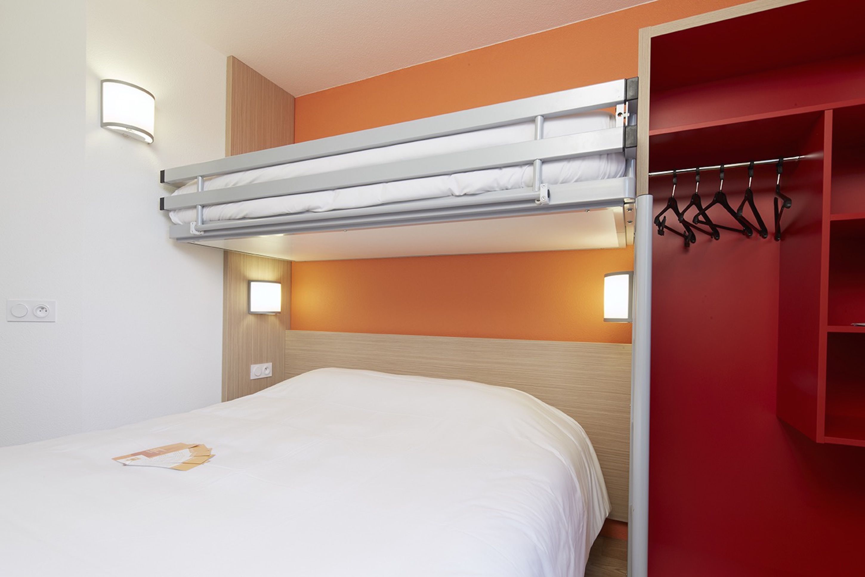 Una cama litera con colchón blanco y una escalera plegable, un cabecero de madera y un armario rojo con perchero.