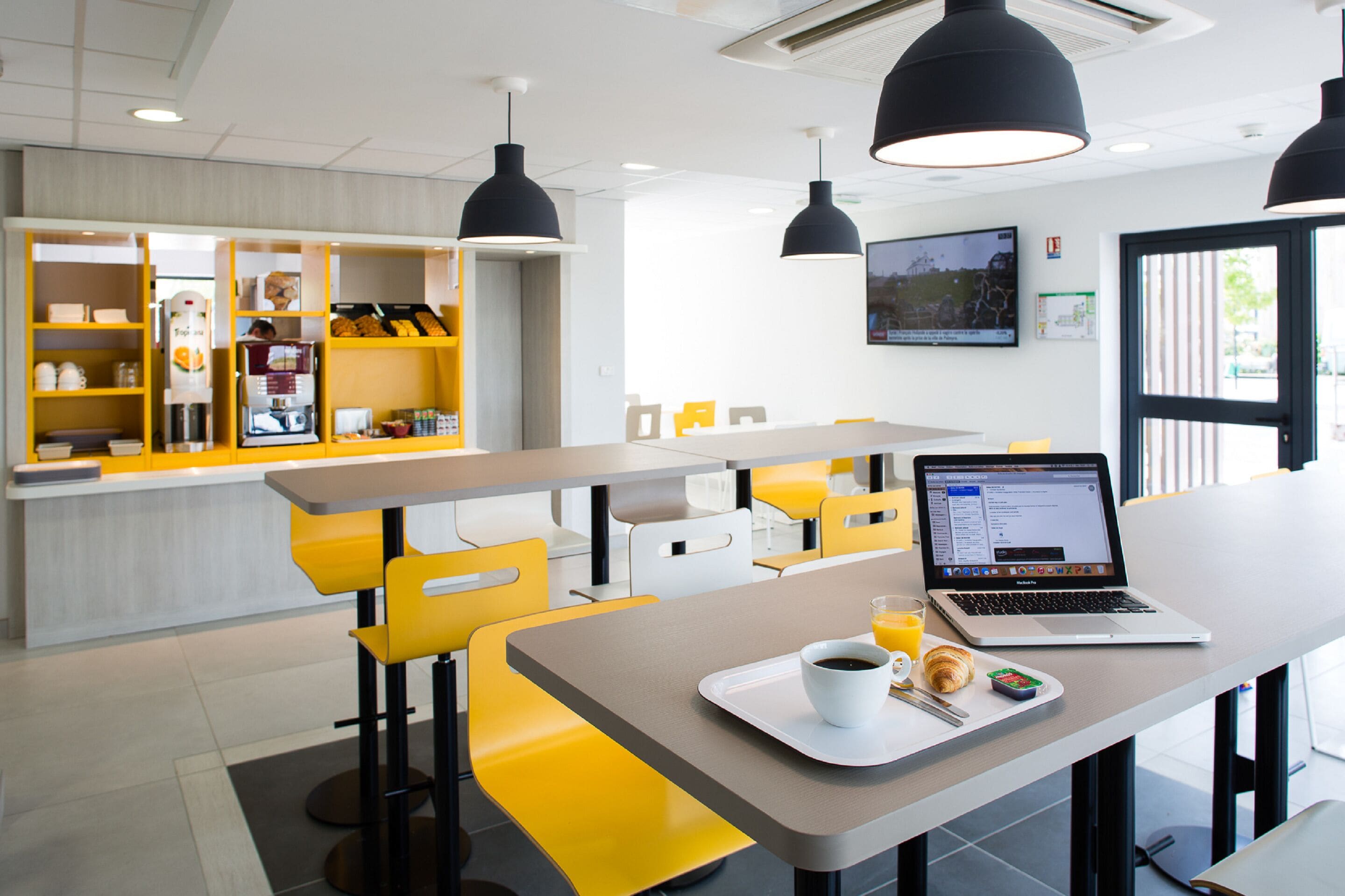 Un café moderno con asientos amarillos y blancos, una máquina de café y un ordenador portátil sobre una mesa.