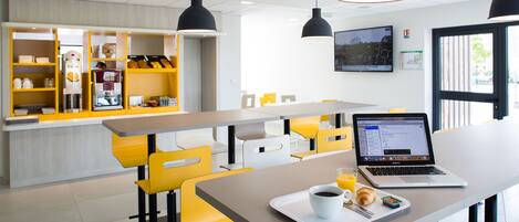 A modern cafe with yellow and white seating, a coffee machine, and a laptop on a table.