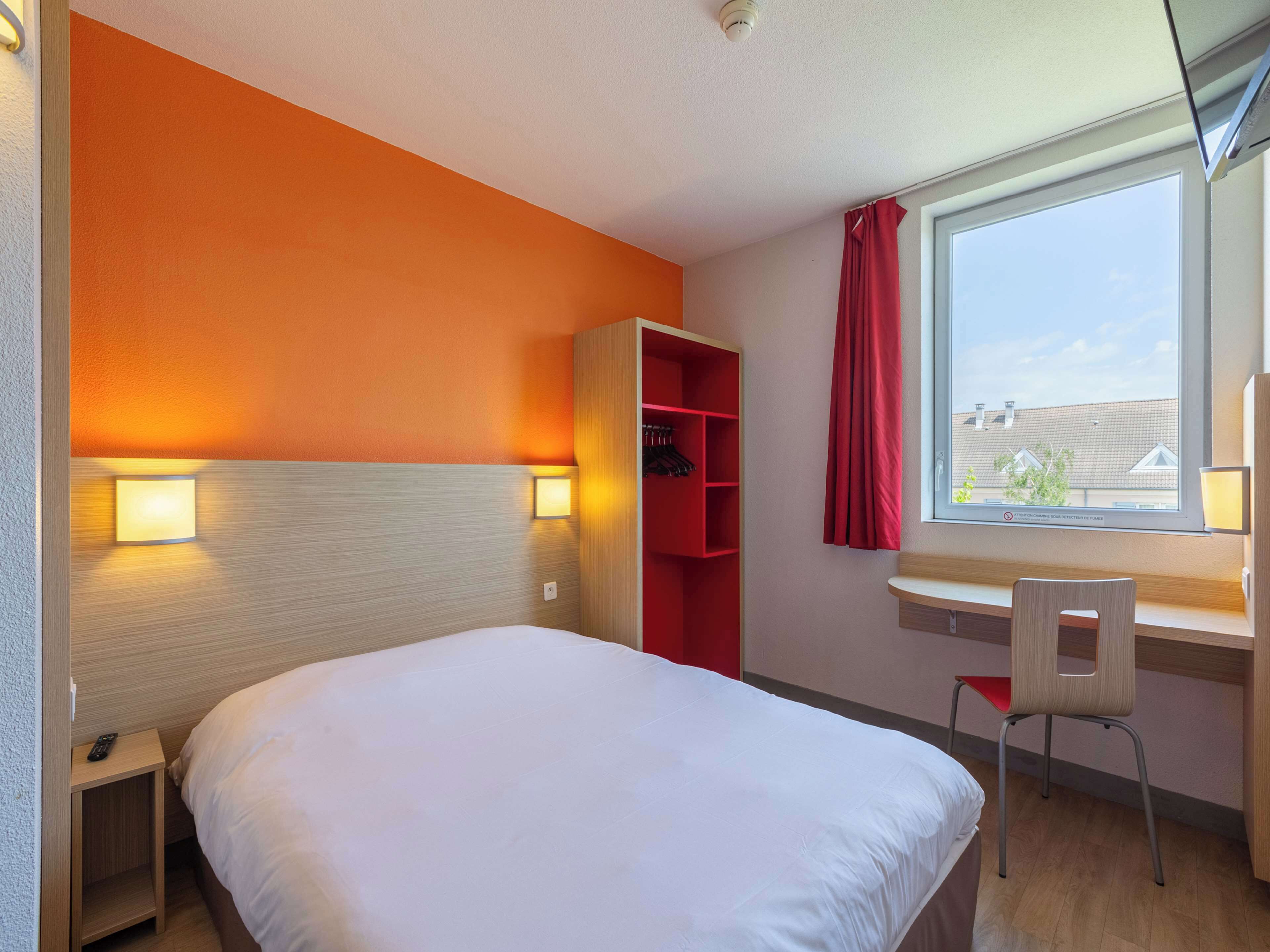 A hotel room with a bed, a desk with a chair, a red wardrobe, and a window with a view of buildings.