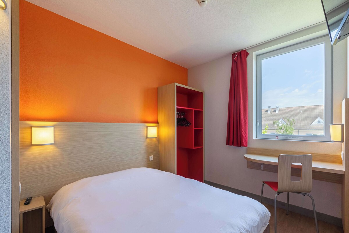 Une chambre d’hôtel avec un lit, un bureau et une chaise, une armoire rouge et une fenêtre donnant sur des immeubles.