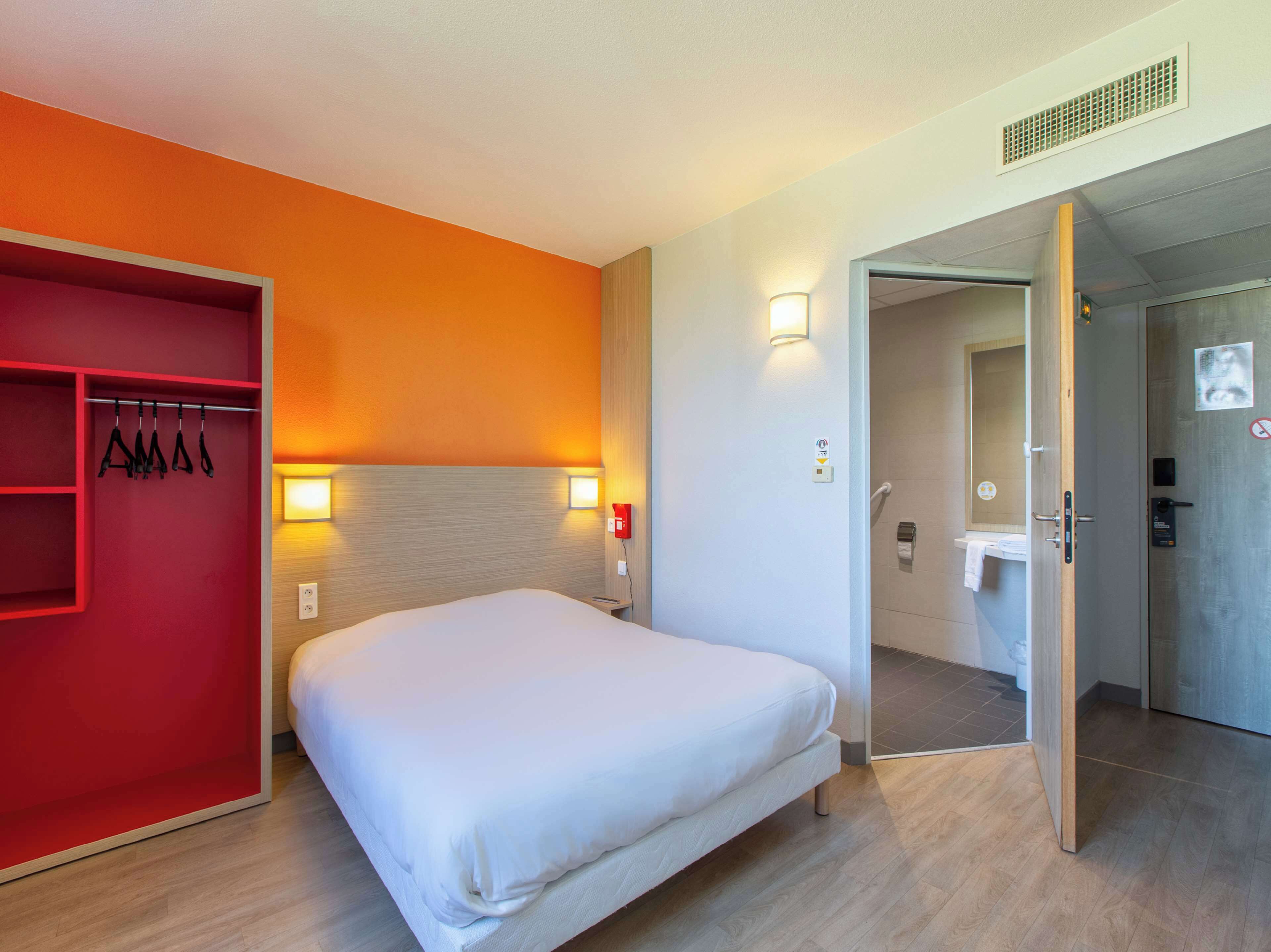 A hotel room with a bed, a red storage unit, and a bathroom visible through an open door.