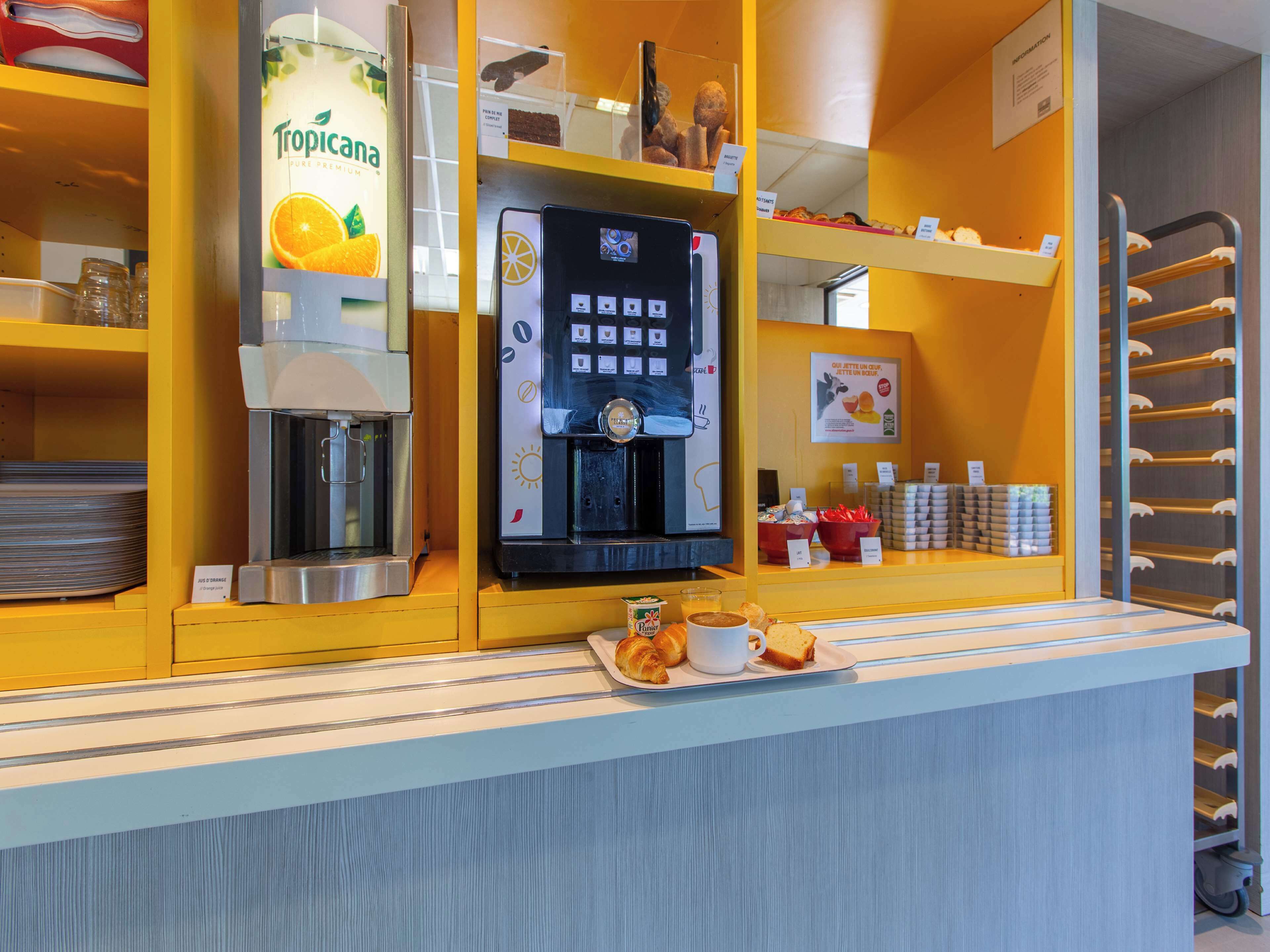 A coffee station with a Tropicana juice dispenser, a coffee machine, and a selection of pastries and bread.