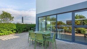 Een buitenzitplaats met tafels en stoelen, een grindpad, en een glazen wand die de omliggende begroeiing en een gebouw op de achtergrond weerspiegelt.