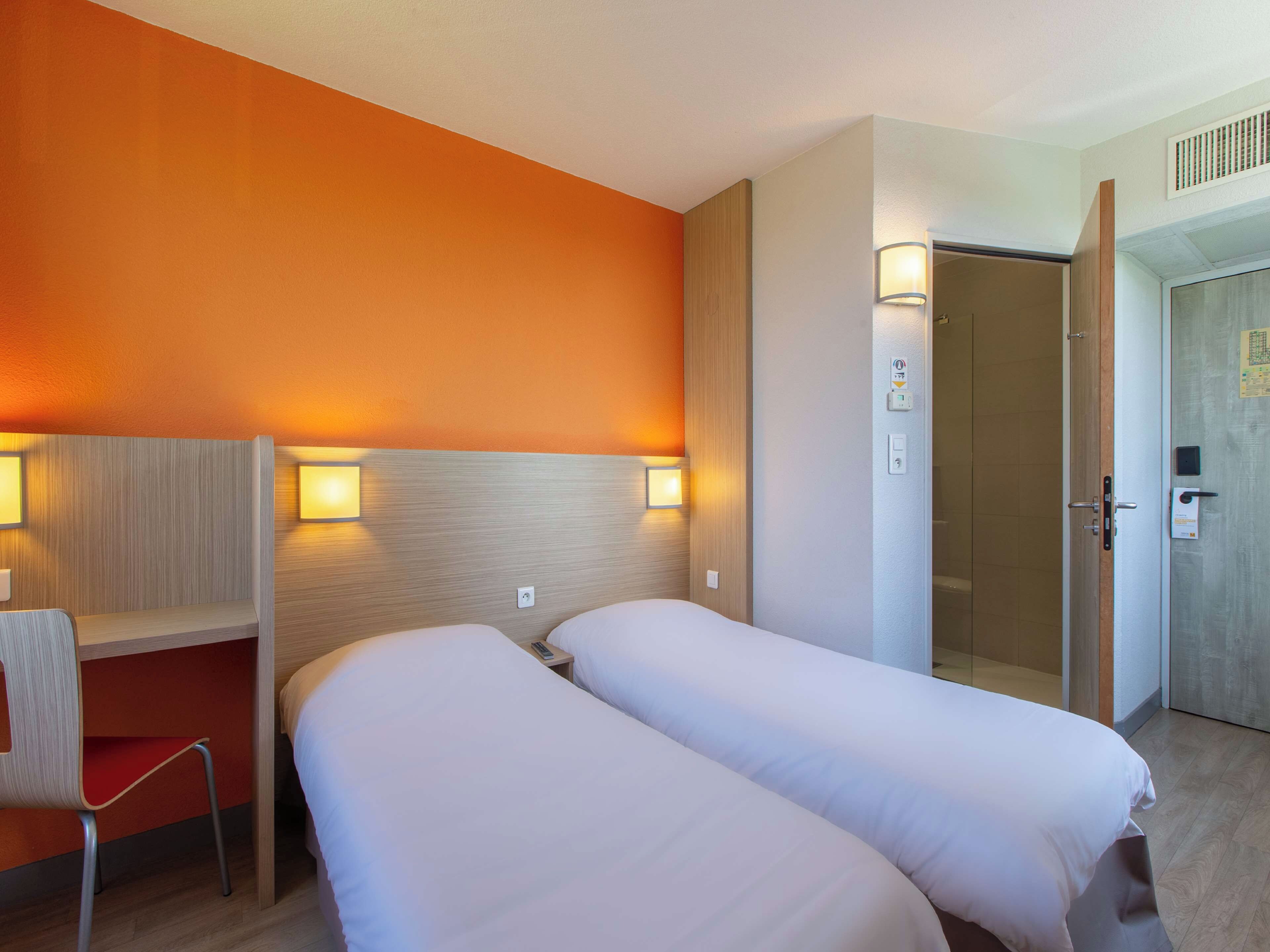 A hotel room with two beds, a desk, and a chair. There is a door to the right and a vent on the wall.