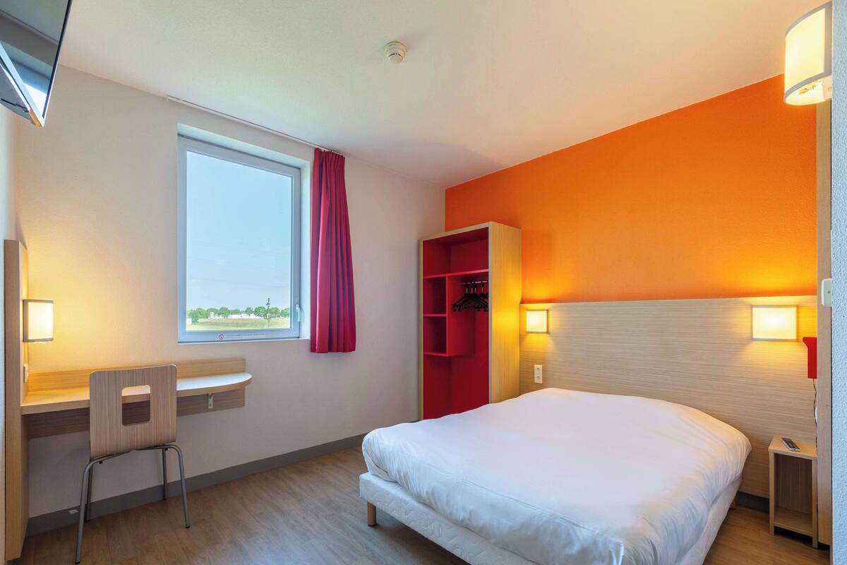 Une chambre d’hôtel comprenant un lit, un bureau, une chaise, une armoire et une fenêtre avec des rideaux.