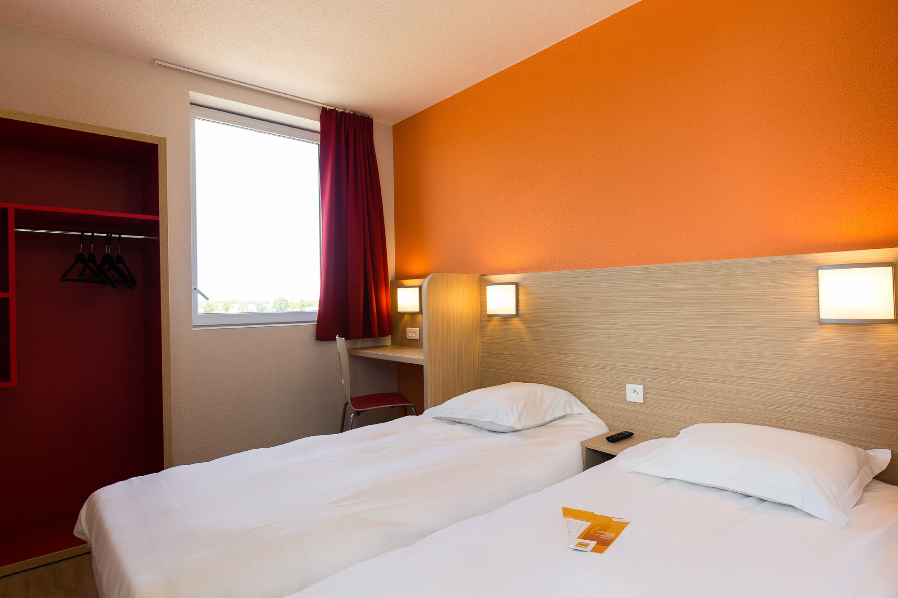 Habitació d'hotel amb dos llits, una paret vermella, una finestra amb cortina vermella i un panell de fusta al capçal.