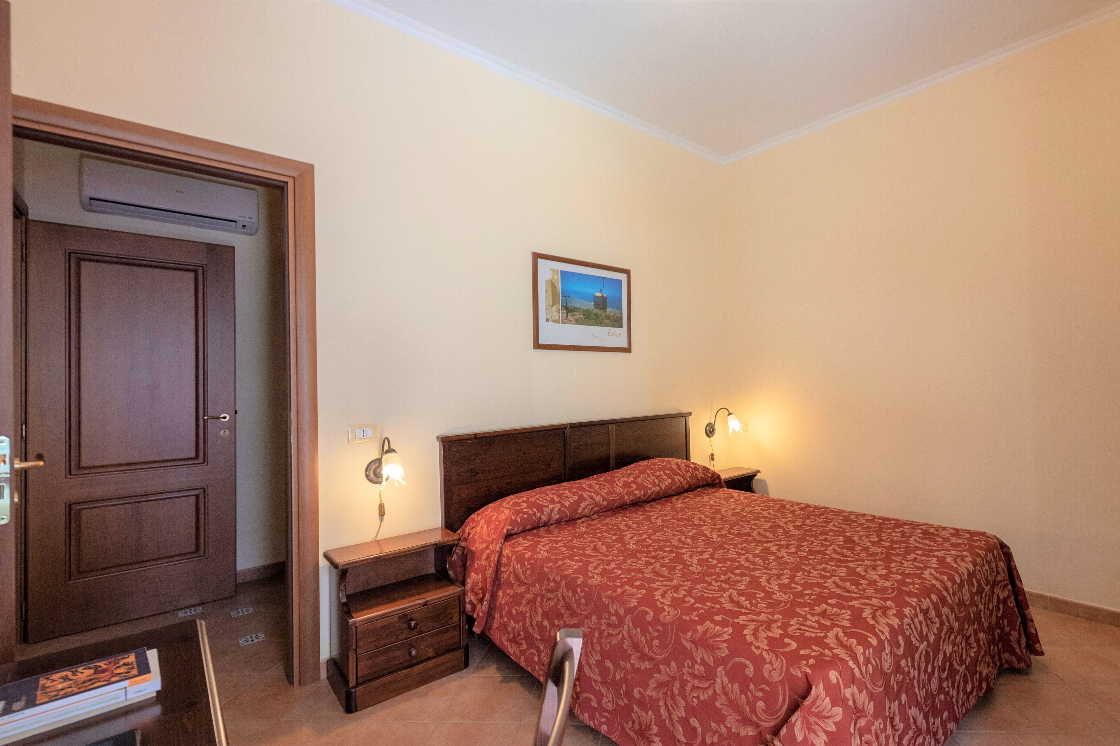 A bedroom with a wooden door, a bedside table with a lamp, a bed with a red patterned bedspread, and a framed picture on the wall.