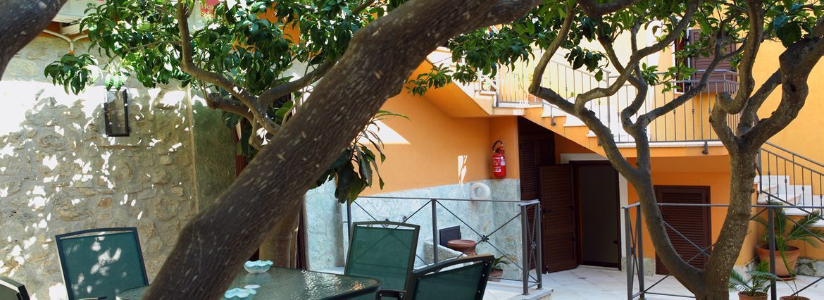 An outdoor seating area with tables and chairs, a stone wall, and a tree providing shade.