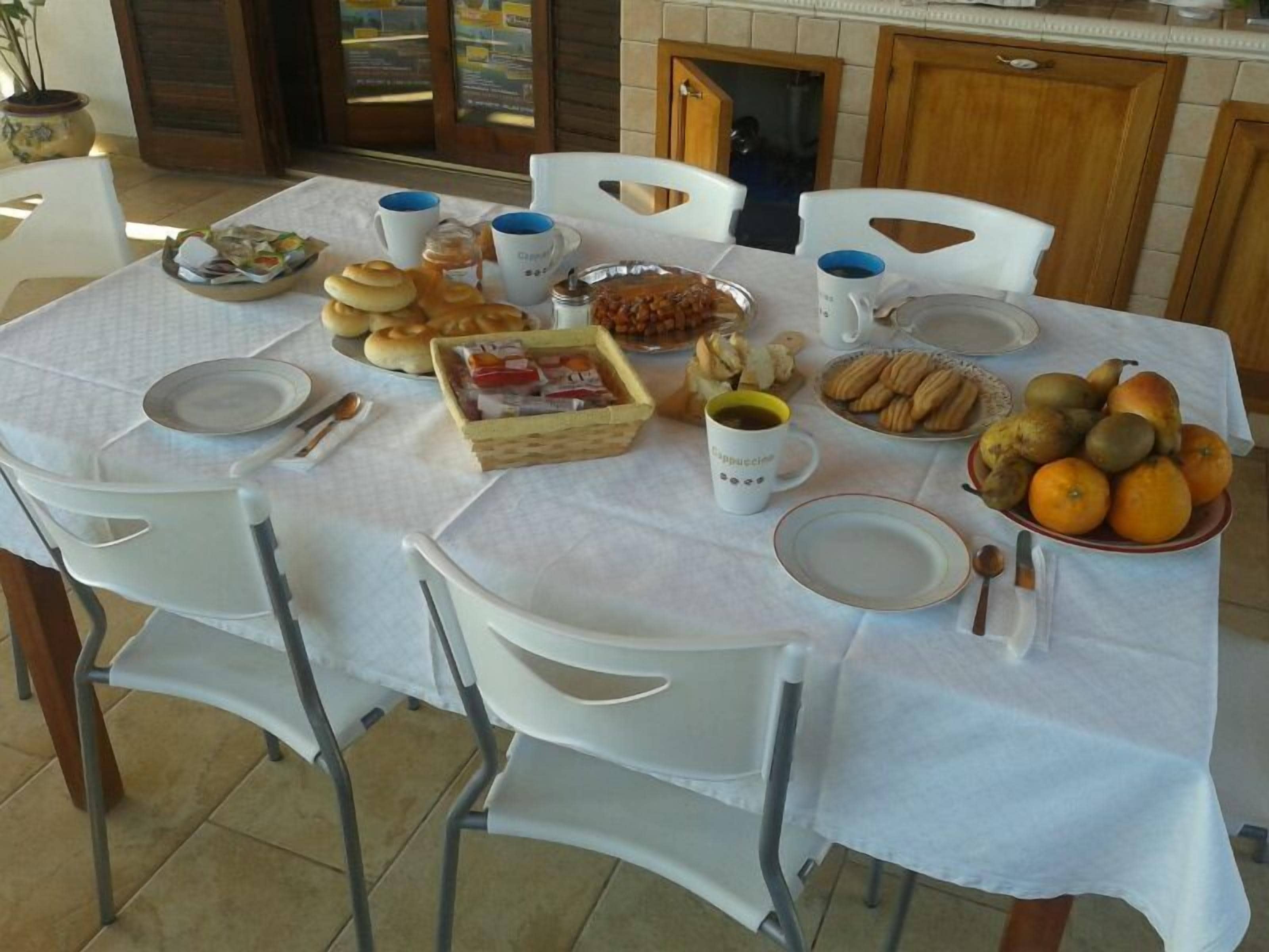 A dining table set with plates, cups, and a variety of food items including pastries, fruits, and a basket of bread.
