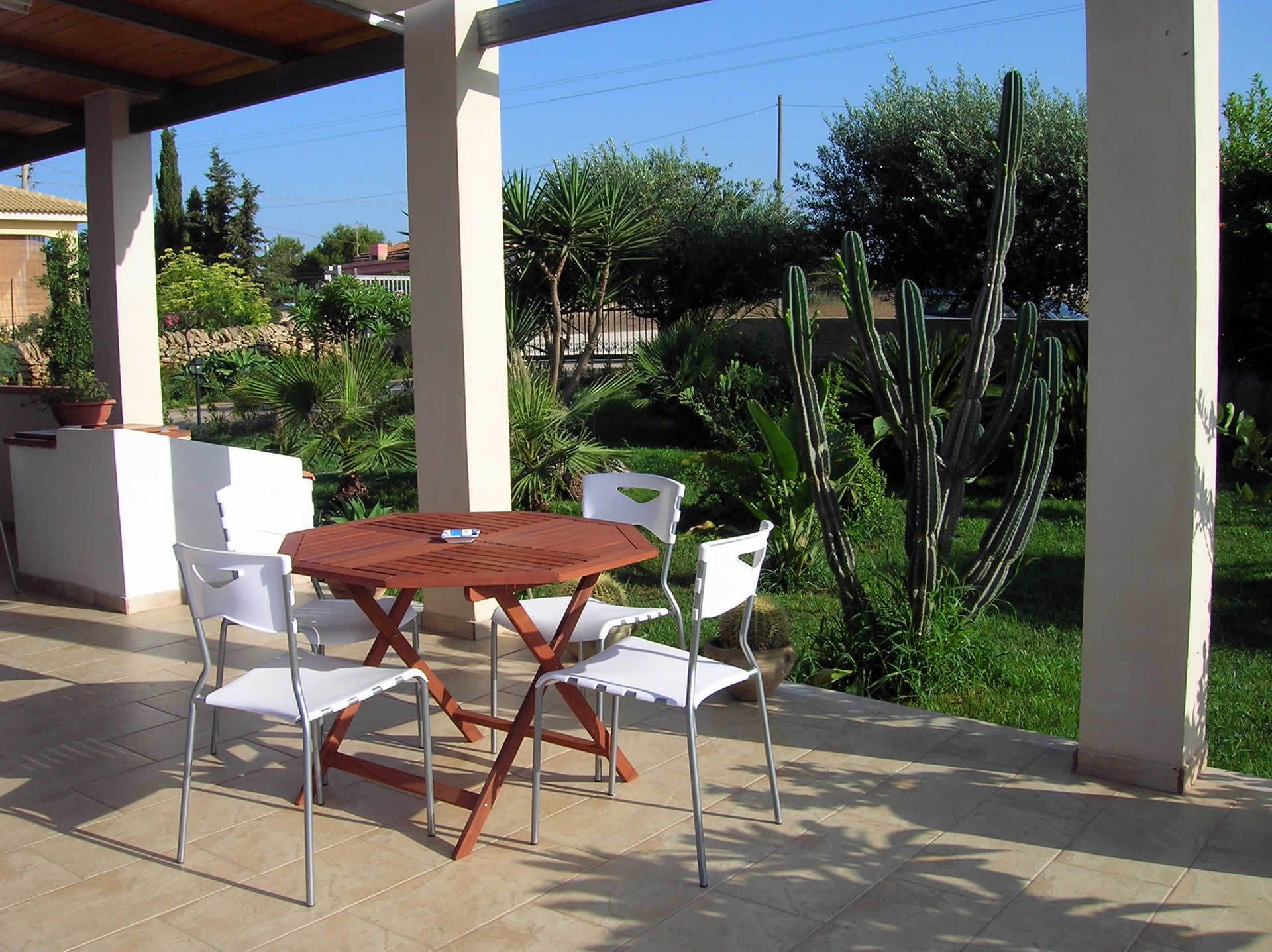 An outdoor patio with a wooden table and white chairs, surrounded by greenery and cacti.