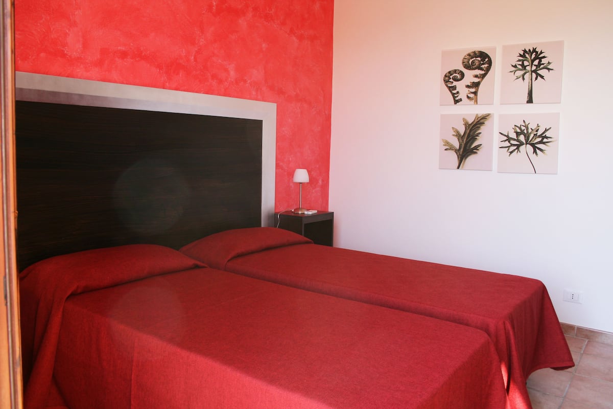 A room with two beds covered in red sheets, a nightstand with a lamp, and four framed pictures on the wall.