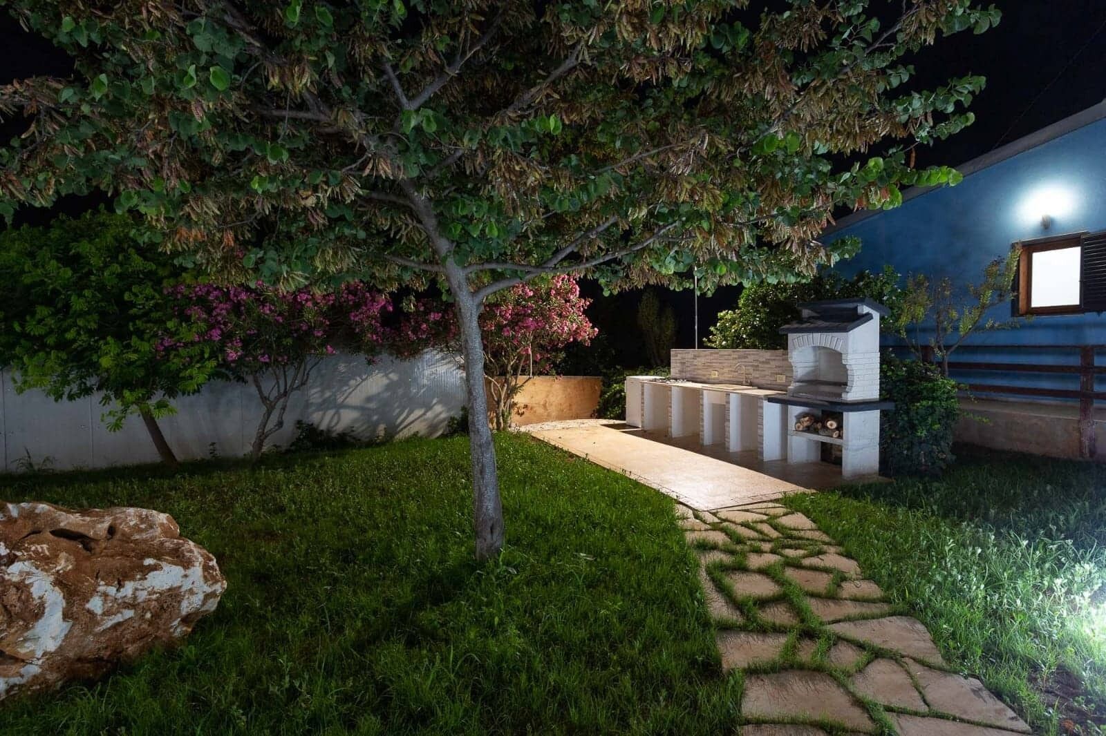 A backyard with a stone pathway leading to a barbecue area, surrounded by greenery and a tree.