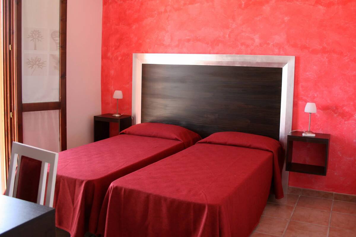A room with two single beds, each with a red bedspread and matching pillows. There are two bedside tables with lamps, and the walls are painted red.