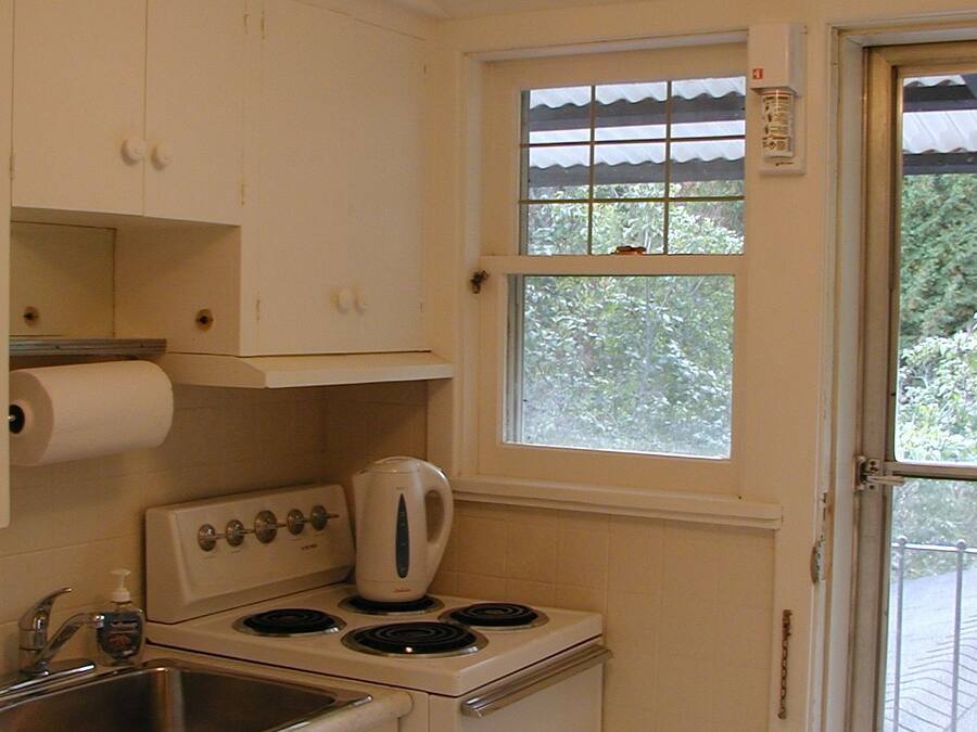 Una cocina pequeña con armarios blancos, estufa, hervidor y ventana con vistas a árboles.