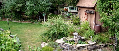 Un jardín con un cobertizo de madera, un comedero para pájaros y una fuente de piedra.