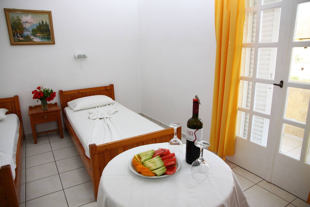 Quarto de hotel com uma cama, mesa de cabeceira com um vaso de flores, prato de frutas, garrafa de vinho e um copo.