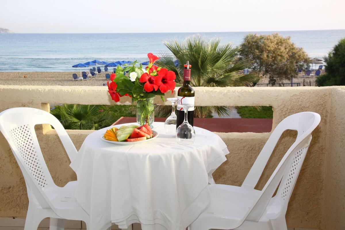 Uma mesa com toalha branca, uma garrafa de vinho, um copo e um prato de frutas numa varanda com vista para a praia.