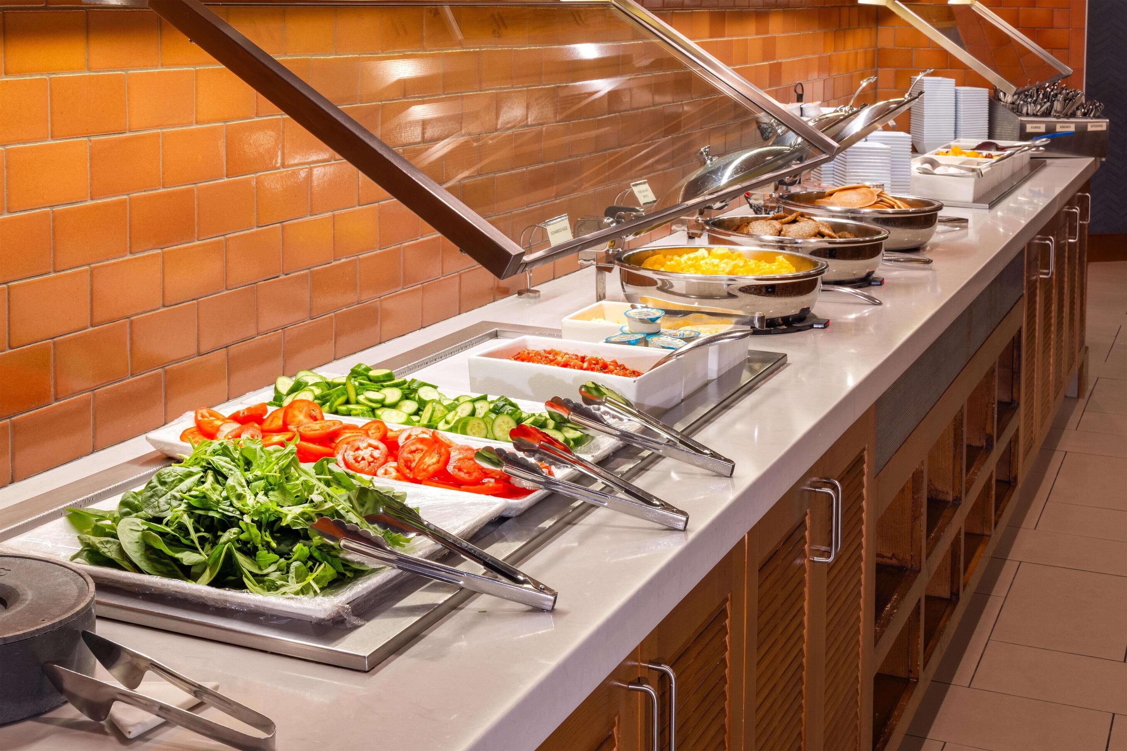 Un bufé con diversos alimentos, como ensaladas y platos calientes, dispuesto en un mostrador largo, con una pared de azulejos al fondo.