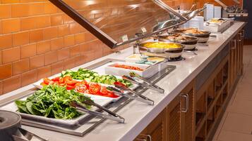 Un bufé con diversos alimentos, como ensaladas y platos calientes, dispuesto en un mostrador largo, con una pared de azulejos al fondo.