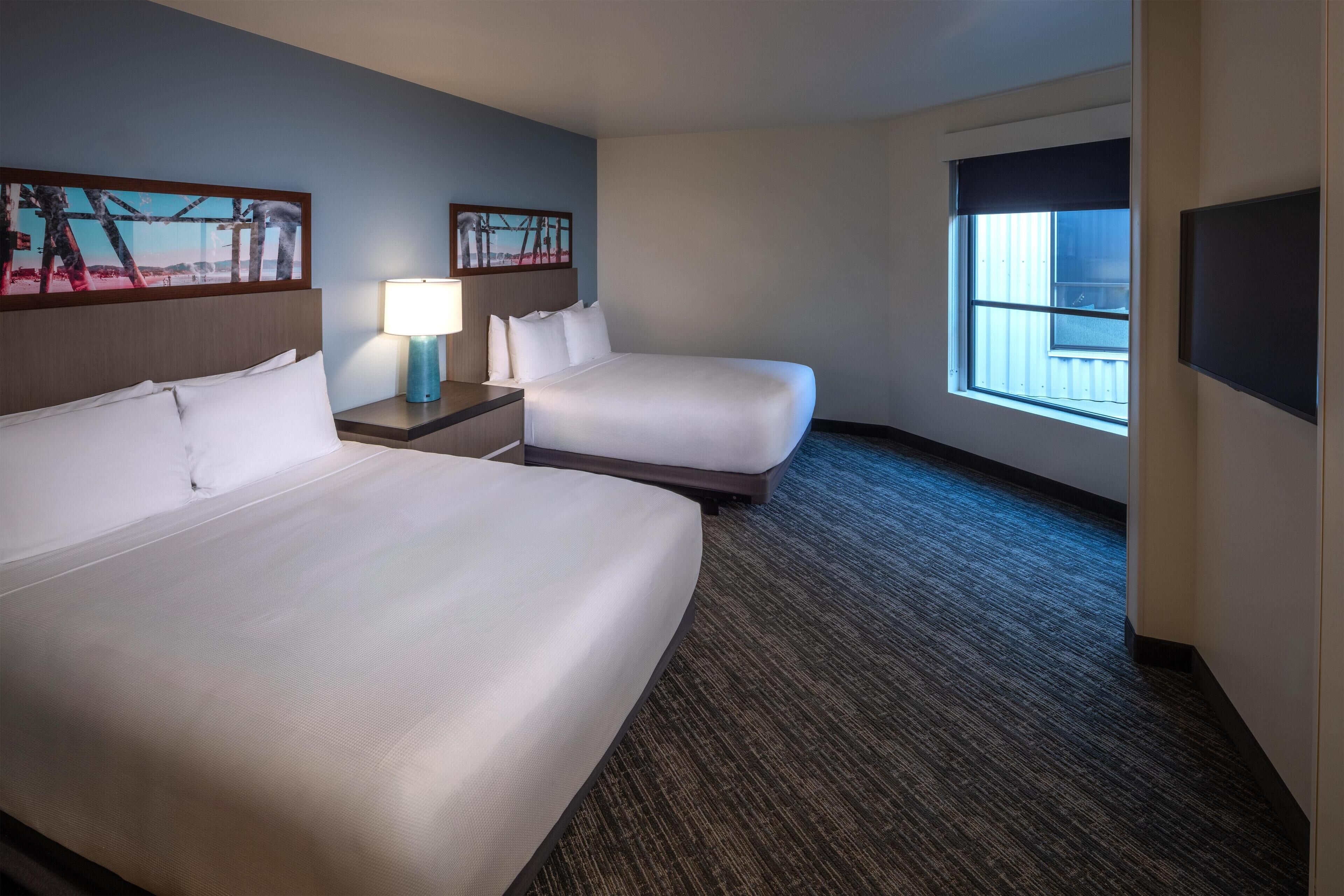 A hotel room with two beds, a nightstand with a lamp, a window with blinds, and artwork on the walls.