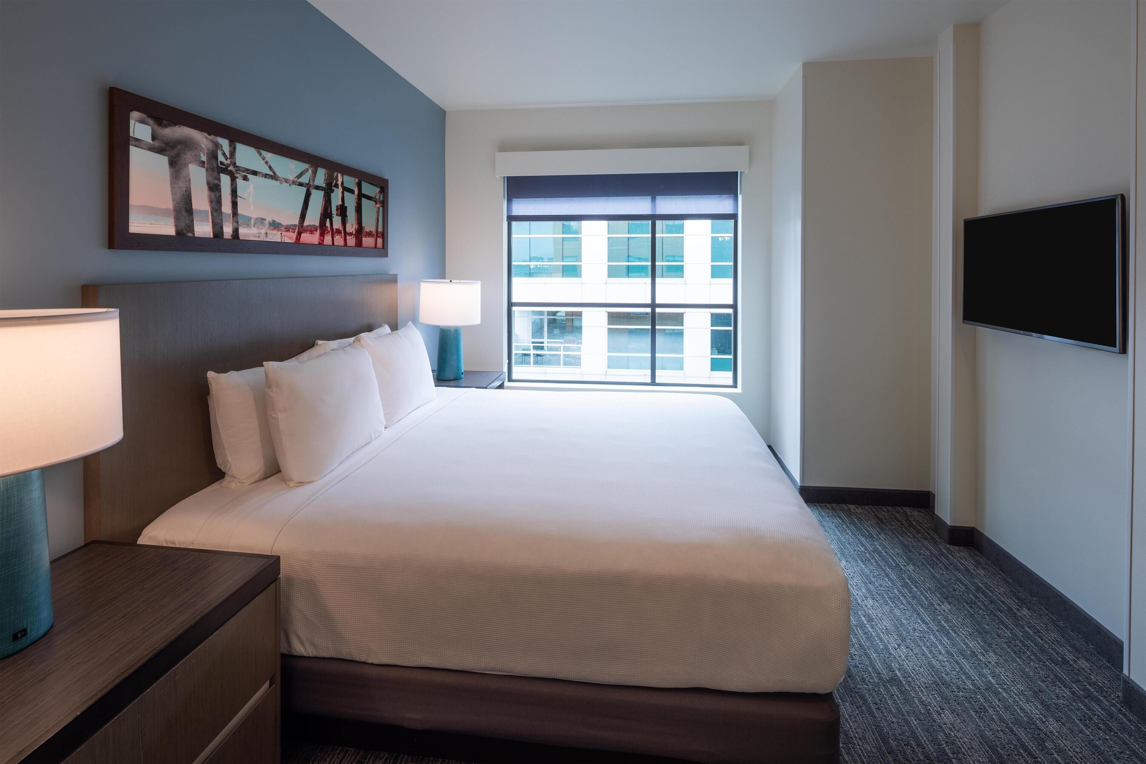 A hotel room with a large bed, a nightstand with a lamp, a flat-screen TV mounted on the wall, and a window with a view of buildings.