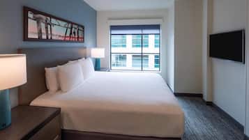 A hotel room with a large bed, a nightstand with a lamp, a flat-screen TV mounted on the wall, and a window with a view of buildings.