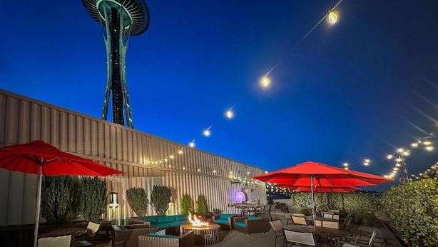 An outdoor patio with seating, a fire feature, and string lights against a backdrop of a tall tower and a clear sky. - Hyatt House Seattle/Downtown (Seattle)