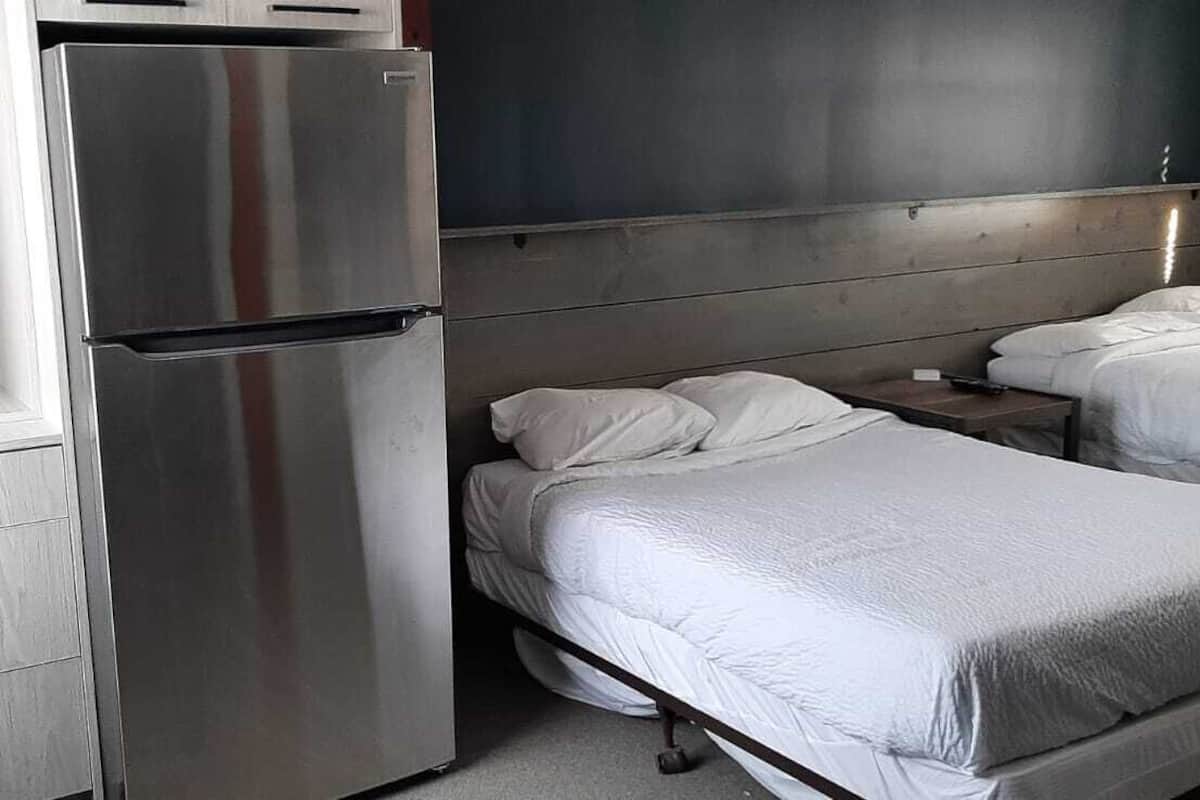 Habitación de hotel con refrigerador metálico, dos camas con ropa blanca, cabecero de madera y una mesita de noche pequeña.