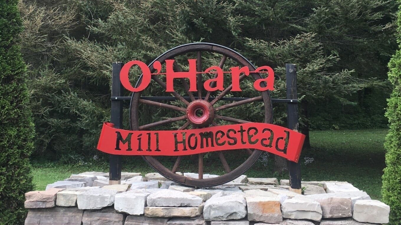 A sign with 'O'Hara Mill Homestead' written on it, mounted on a stone base with a wooden wheel structure.