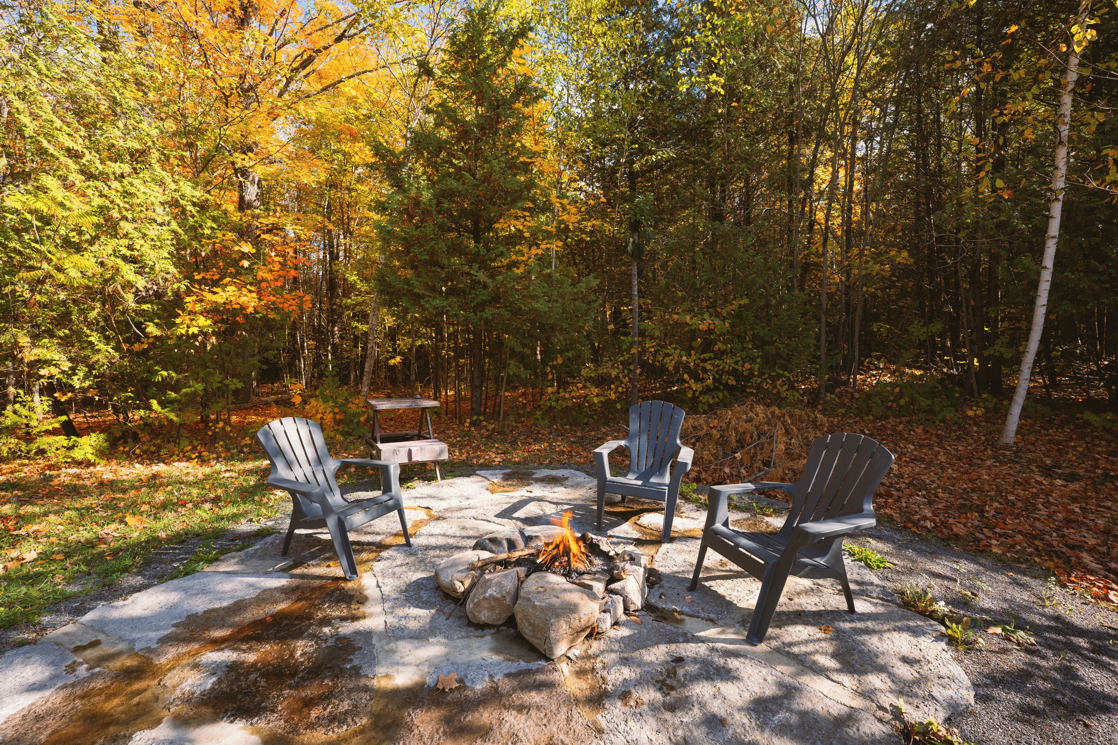 A campsite with a fire pit, three Adirondack chairs, and a wooden table.