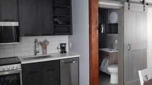 A modern kitchen with dark cabinets, a stove, and a dishwasher. A dining area with a table and chairs is visible. - Moira Lake Motel (Madoc)