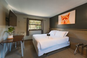 A hotel room with a bed, a desk, a potted plant, a window, and a wall-mounted artwork.