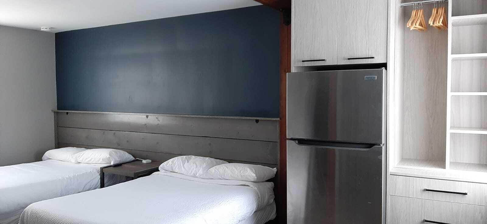 A hotel room with two single beds, a large dark-colored wall, a stainless steel refrigerator, and a wooden cabinet with hanging clothes.
