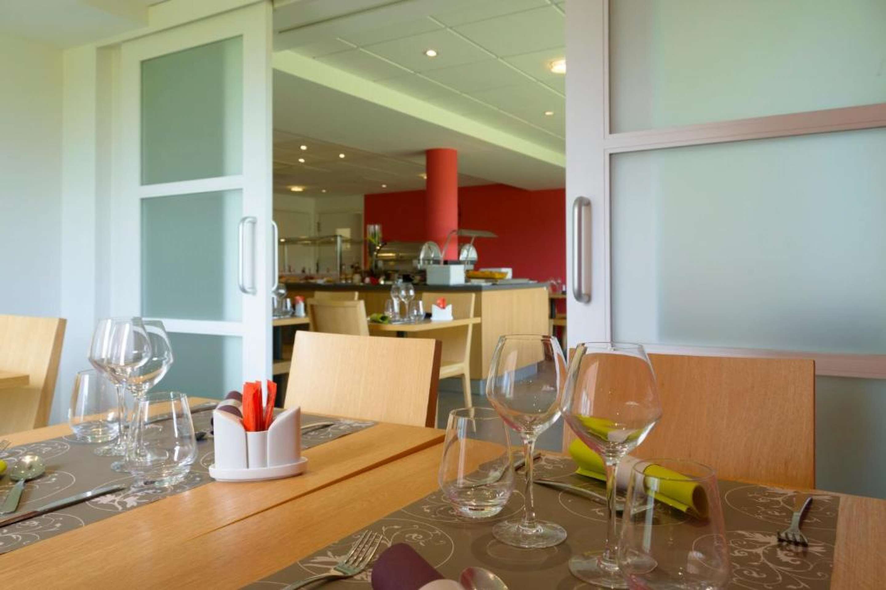A dining area with a wooden table set for two, clear glassware, and a white napkin holder with utensils.