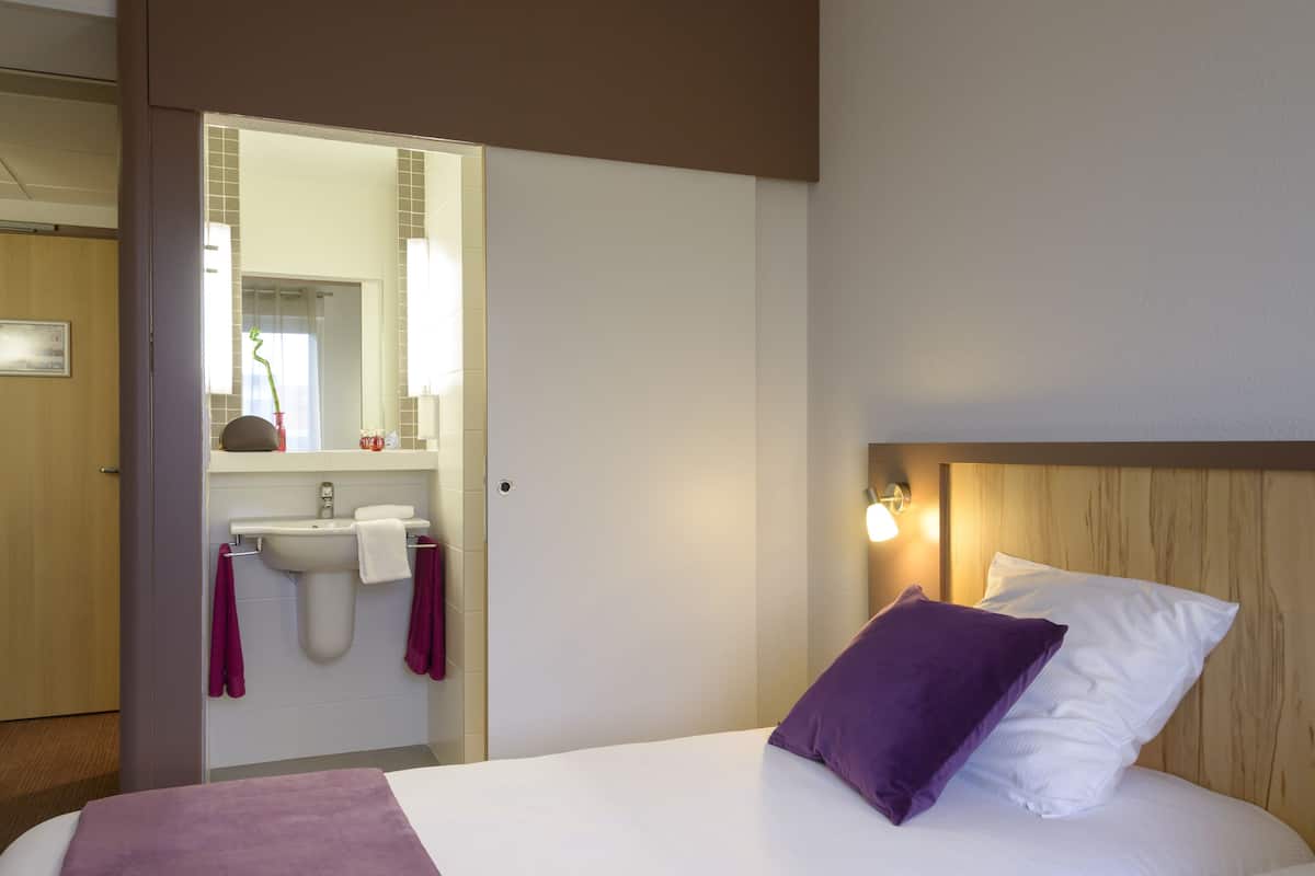 Ein Hotelzimmer mit einem Bett, einem Holz-Kopfbrett, einem Nachttisch, einem Spiegel, einem Waschbecken und einer Tür zu einem weiteren Raum.