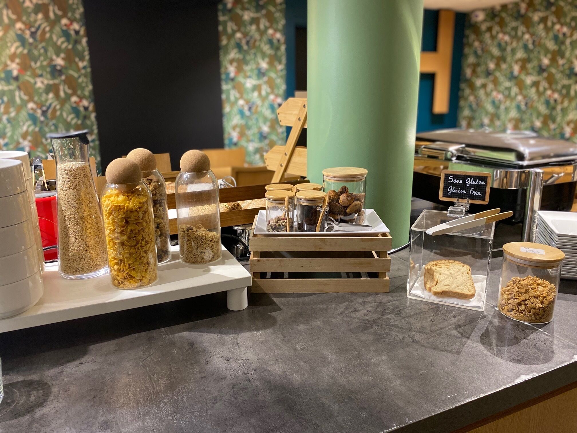 A breakfast buffet with various food items and containers, including jars of granola and nuts, on a counter with a sign indicating gluten-free options.